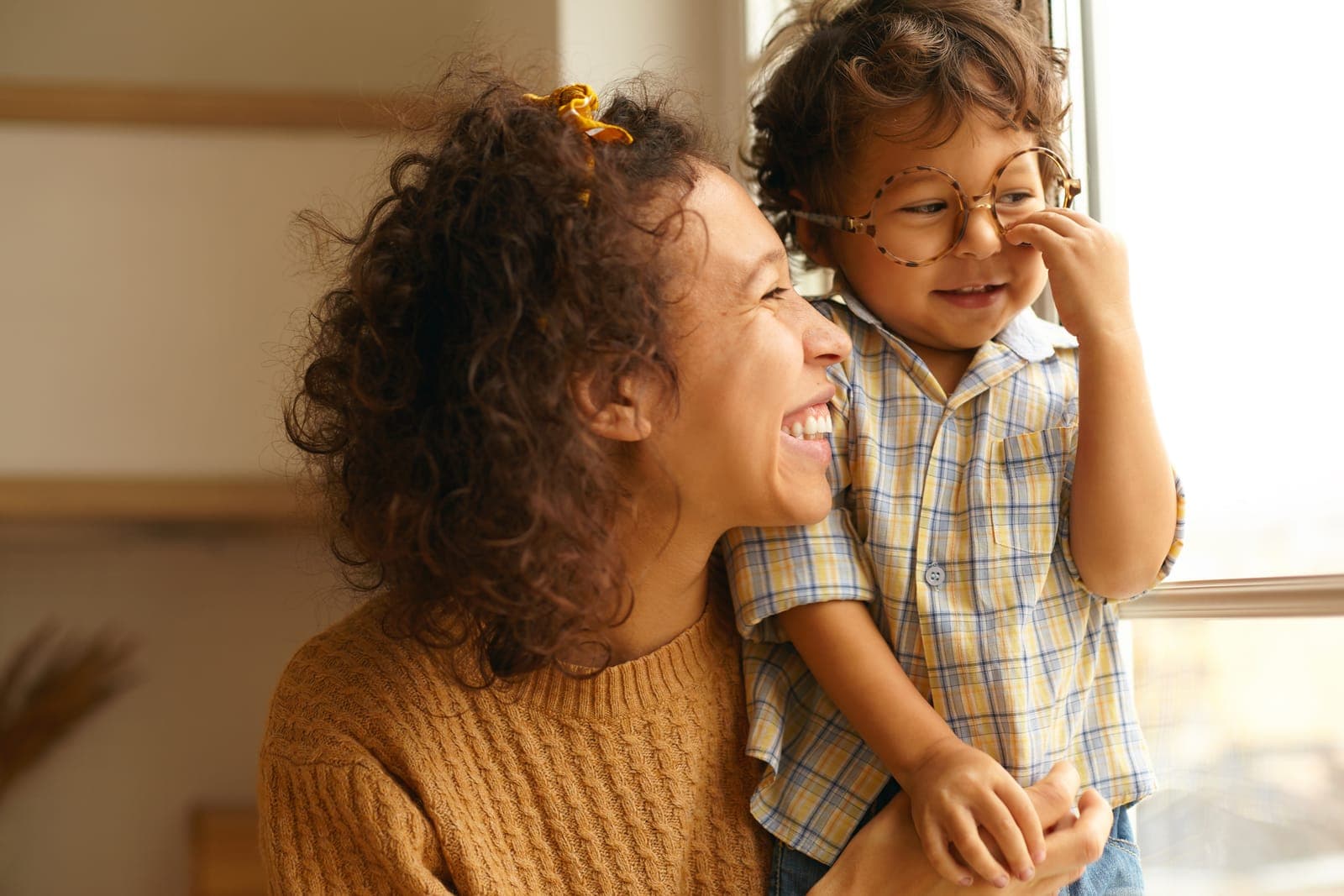 Mãe rindo com o filho no colo perto da janela, sem pressa